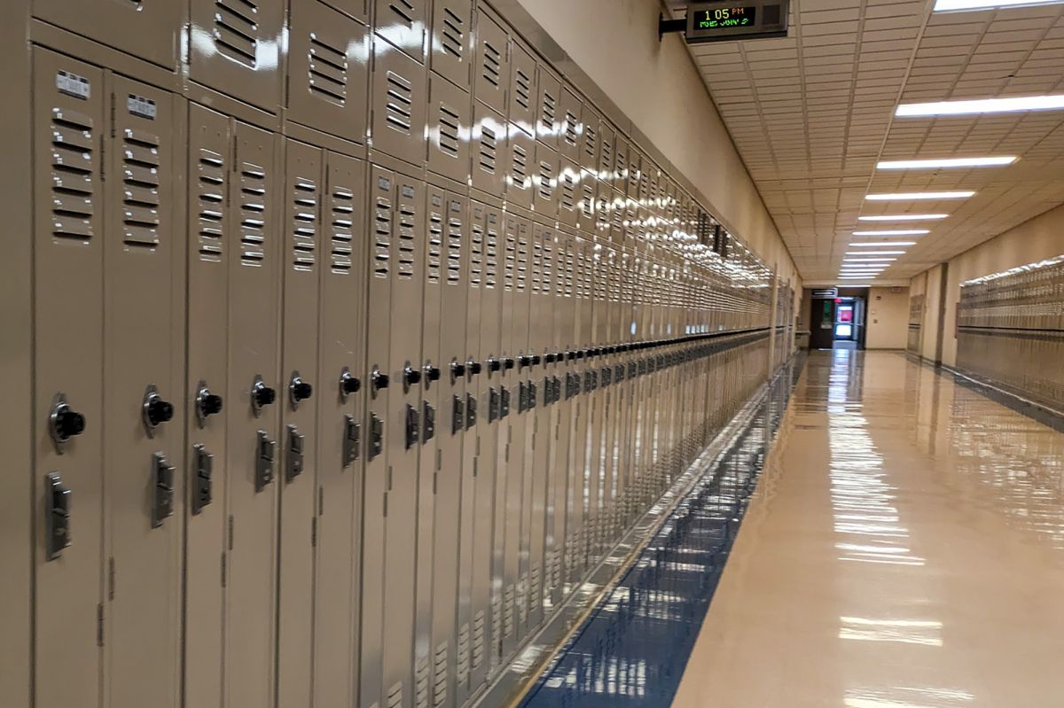 An empty school hallway is lined by lockers. A digital clock at the top of the hallway reads the time and date. The fluorescent lights running down the hallway reflect off the linoleum flooring.