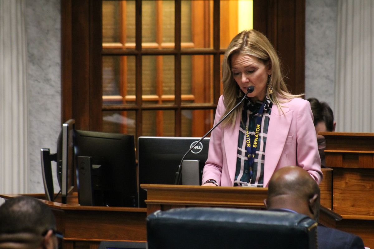 Shelli Yoder wears a pink blazer and speaks into a microphone. She is a White woman with shoulder-length blonde hair.