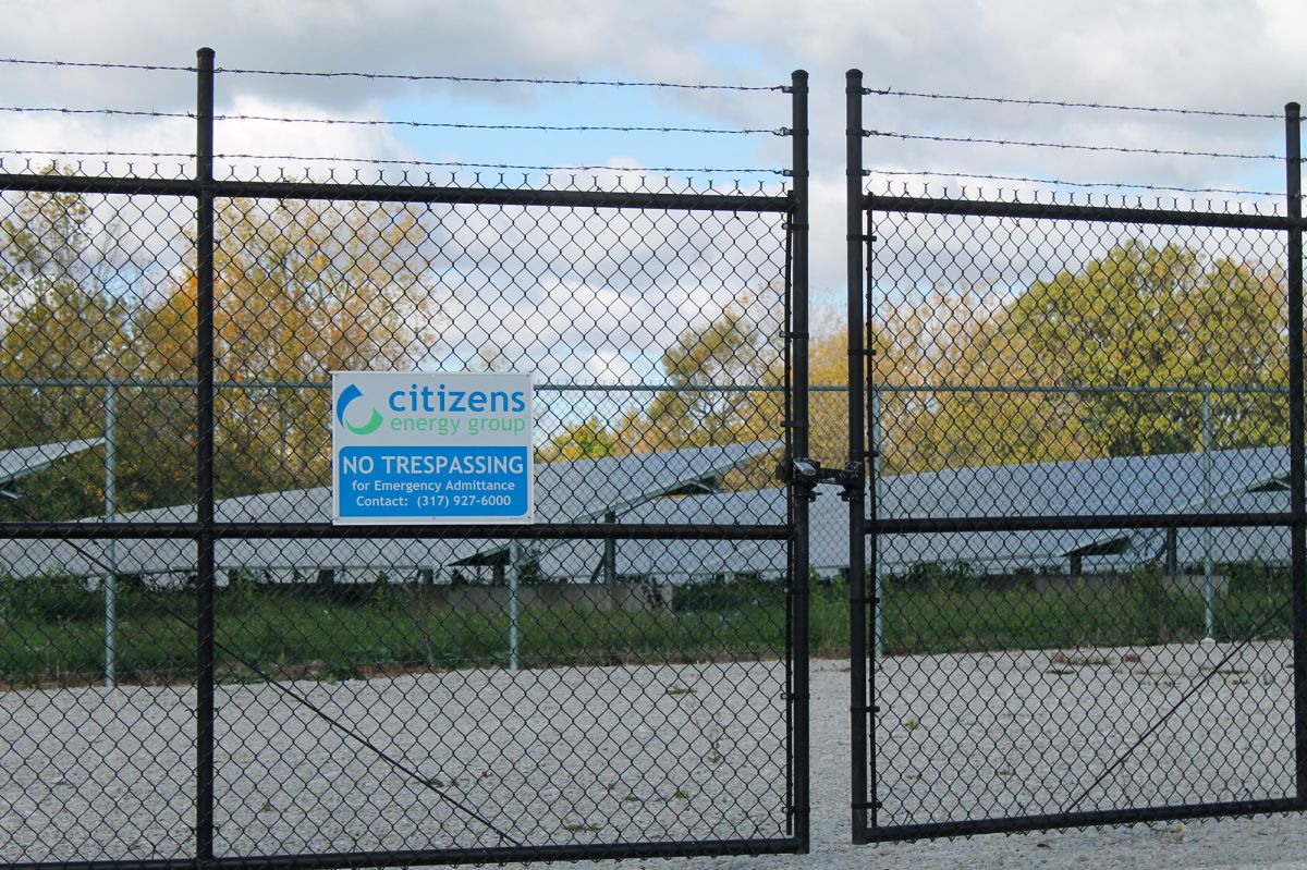 A Citizens Energy solar farm in Indianapolis. Solar panels sit behind a black, chainlink fence. A sign is posted on the gate with Citizens's logo above a no trespassing sign. Gravel leads up to a second metal fence before reaching the solar panels an