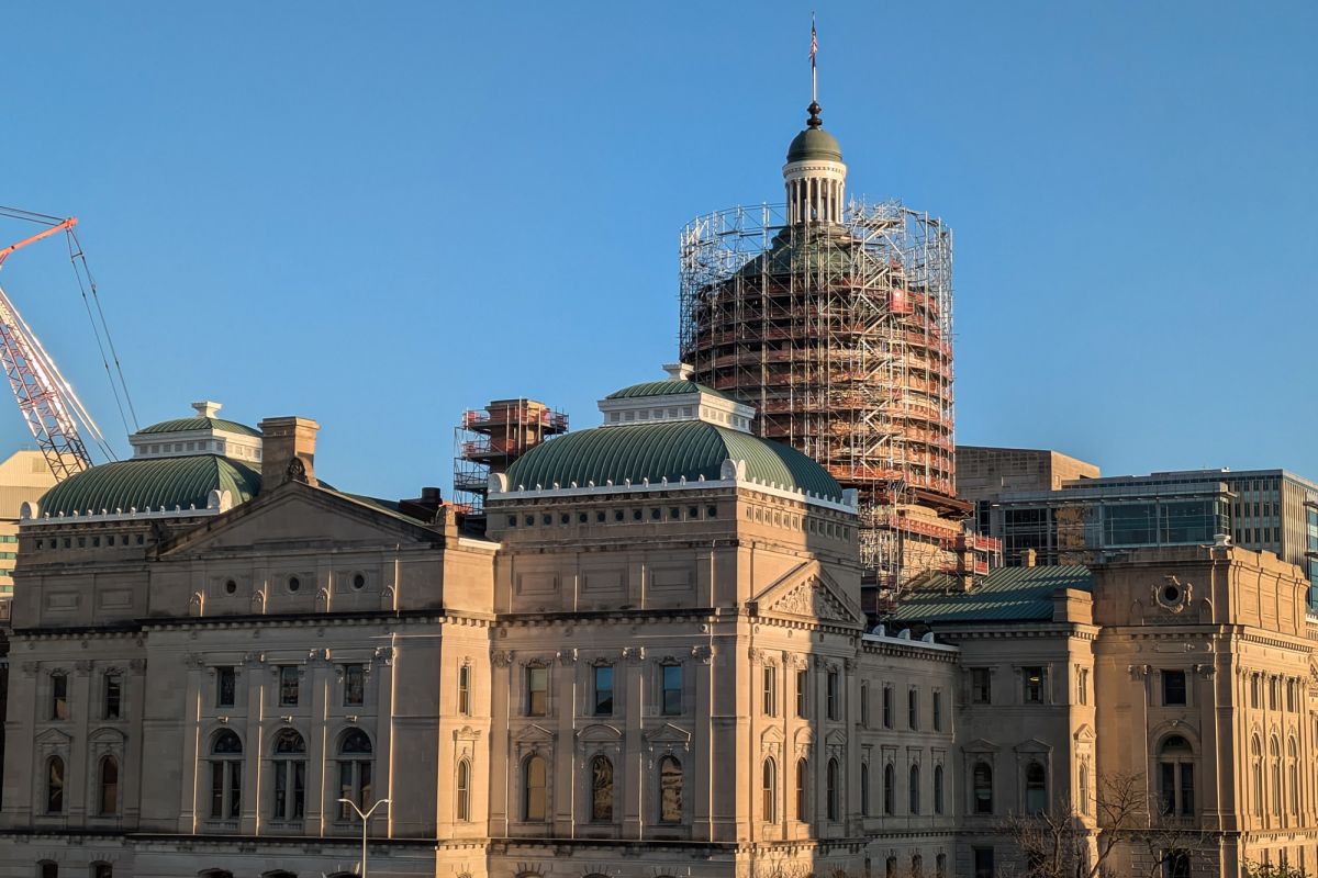 The Indiana Statehouse as seen at sunset from a parking garage at the northwest corner of the building. Scaffolding surrounds the building's dome and a crane is cut off on the far left of the image. Behind the building, the sky is clear and blue.