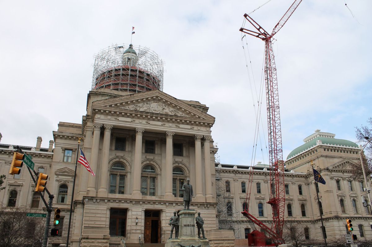 The North Capitol Avenue entrance to the Indiana Statehouse is framed by a stoplight and American flag on one side and a large red crane on the other, with scaffolding surrounding the cupola.