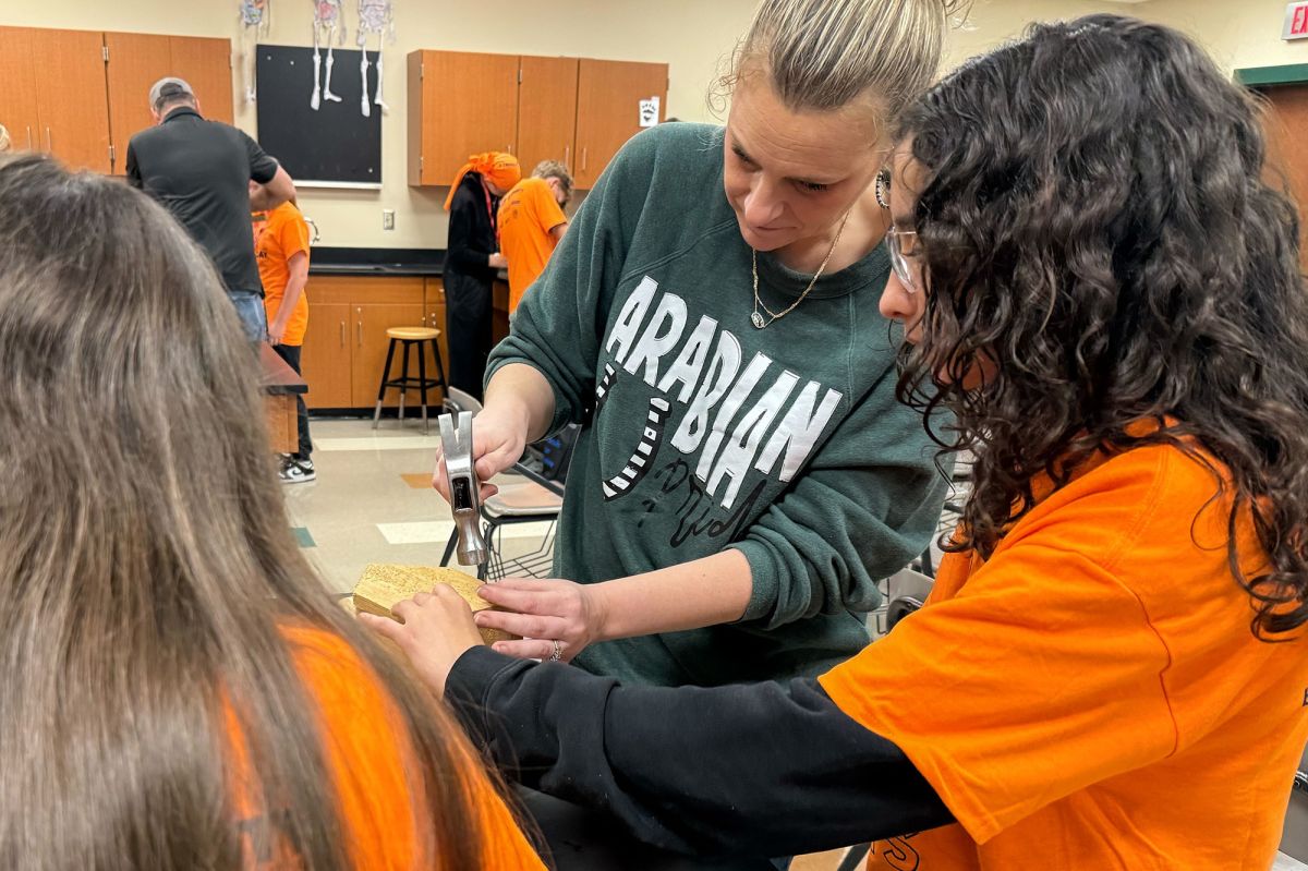 Students wear orange shirts and safety goggles. A woman stands next to the students and wears a green shirt holding a hammer. 