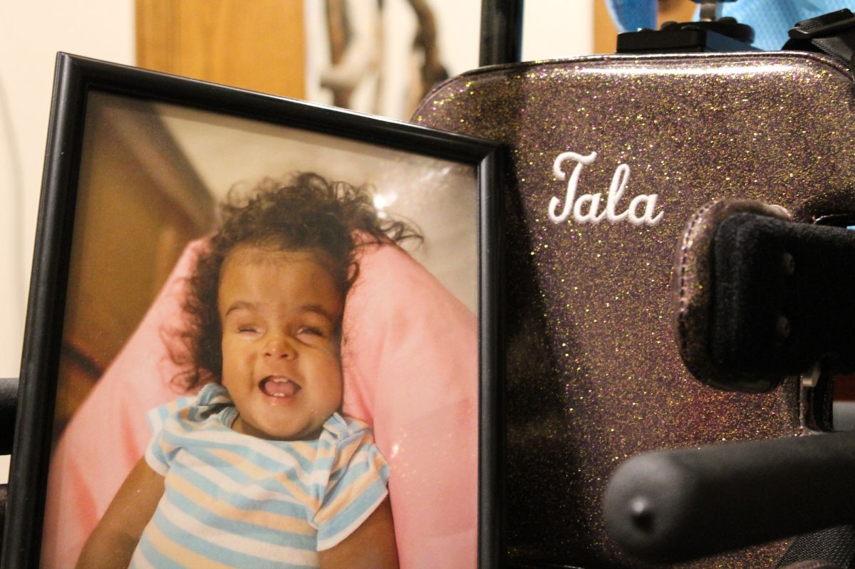 A photo of Tala Beth Davis with a wide smile rests on her wheel chair. The back rest of the chair is sparkly and has the name “Tala” embroidered on it.