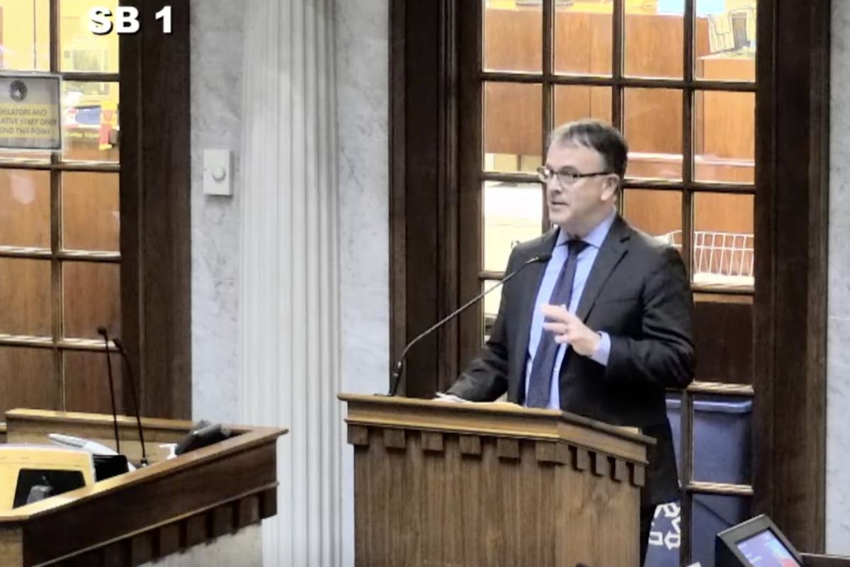 Terry Spradlin raises his hand while standing behind a podium and speaking to lawmakers in the Senate Chamber.