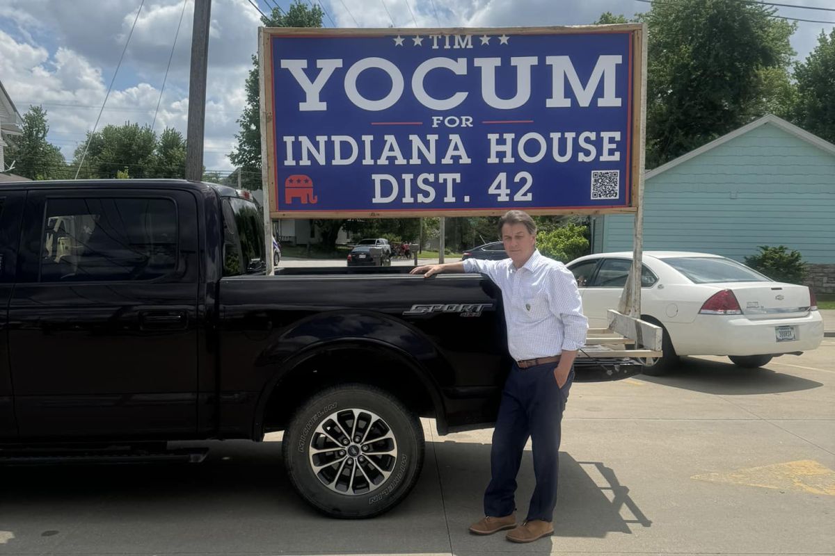 Tim Yocum stands in front of a truck with a sign standing in the truck bed that reads "Tim Yocum For Indiana House Dist. 42". Yocum is a White man with dark hair. He is wearing a white shirt and black pants.