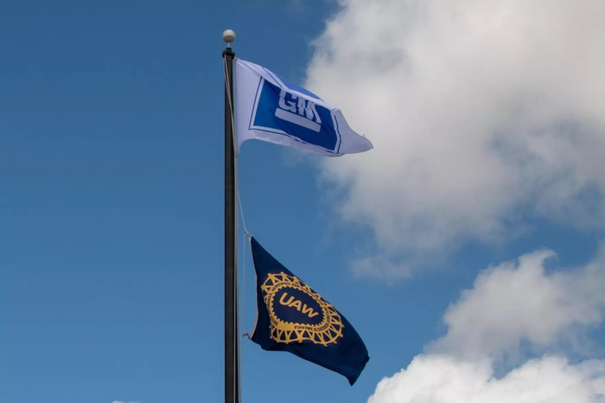 There are two flags on a tall black pole. The first flag has a white base with a blue square in the center with the letters "GM" in a bold white font. Underneath this flag is a black flag that has a gold circle with the words "UAW" in the center" 