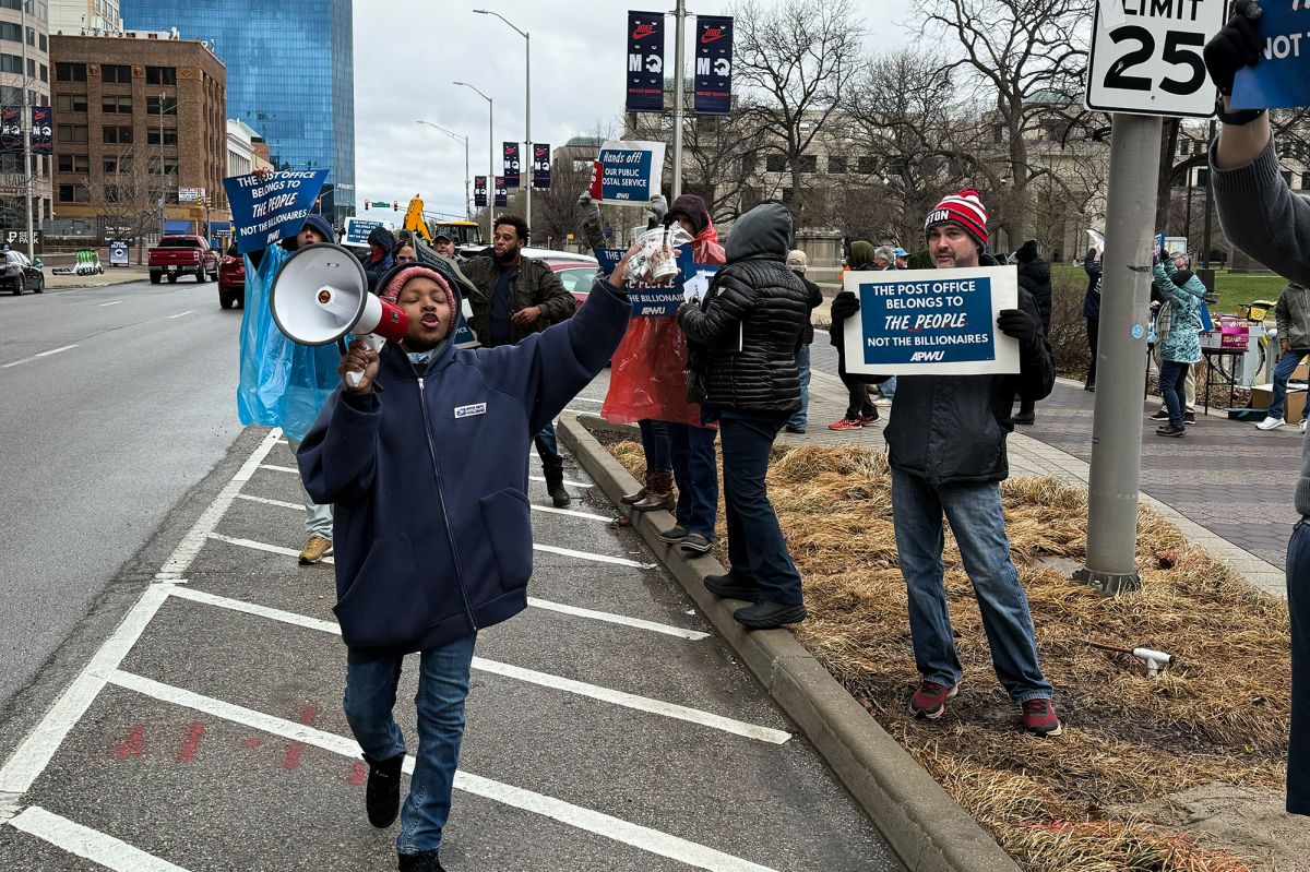 A Black woman holds a white mega phone as she marches down the street in front of the Indiana Statehouse. There are people gathered with signs. 