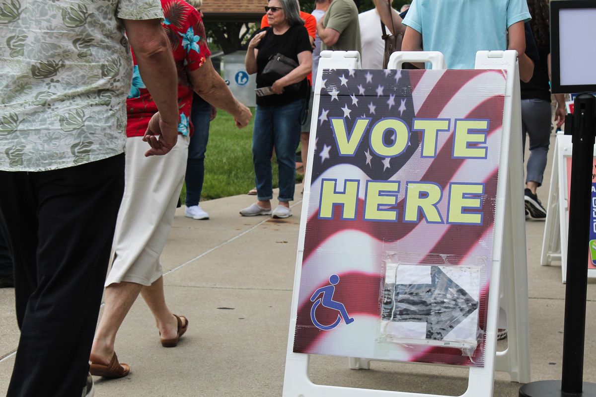 A sidewalk sign reads "Vote Here" with an arrow pointing to the right and a handicap accessibility logo. There are people walking by the sign.