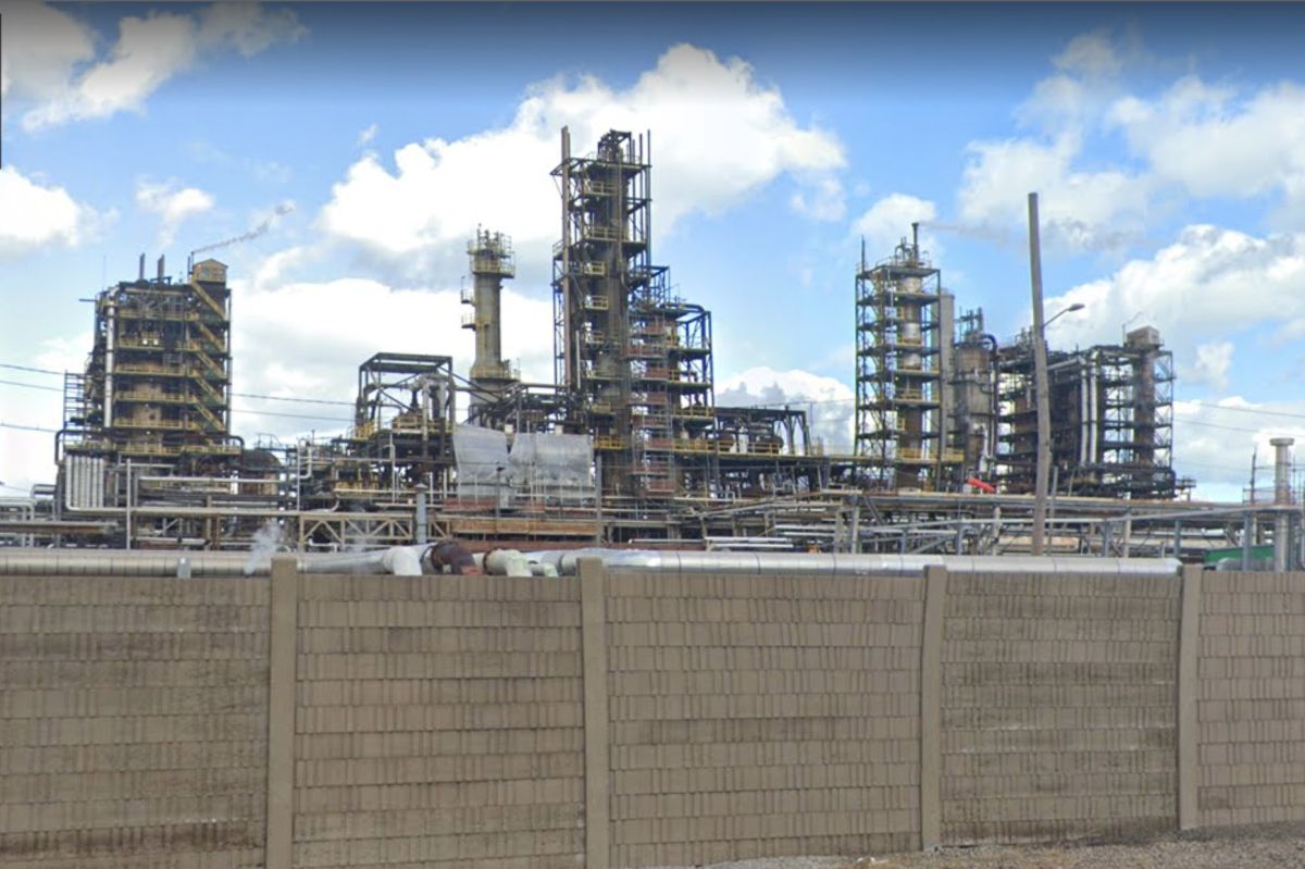 The exterior of BP's Whiting Refinery sits behind a tan brick wall. The Google Map screenshot has blue sky and fluffy clouds behind the industrial refinery.