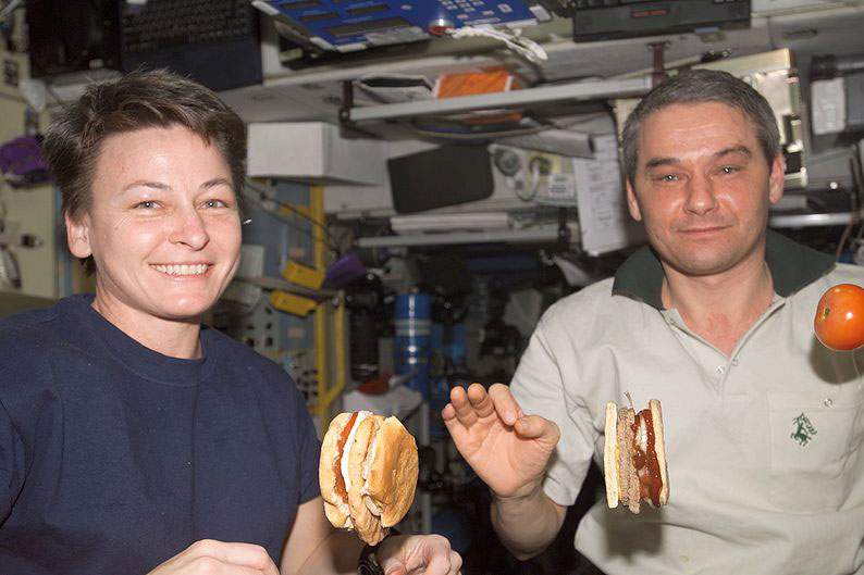 two astronauts sit while their burgers float in the air