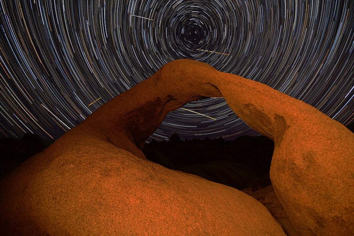 Time-lapse photo of a natural arch at night with a few meteors streaking in the starry sky behind