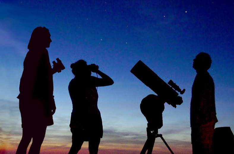 Three people look into the night sky with binoculars and a telescope during a meteor shower.
