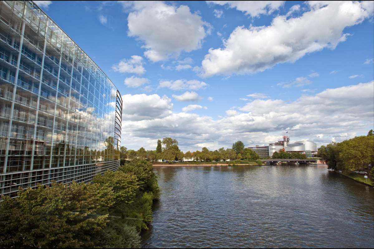 Clouds floating in the sky. The clouds are reflected on the European Parliament building on the left side of the screen. The clouds are reflected by a lake on the right side of the screen.