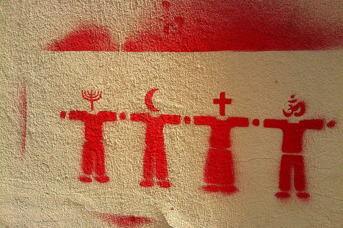 A stenciled image spray painted red on a cream colored wall. The stencil depicts 4 religious symbols as people's heads. The symbols from left to right are the Jewish Menorah, the crescent moon for Islam, the cross for Christianity, and the Om for Hinduism.