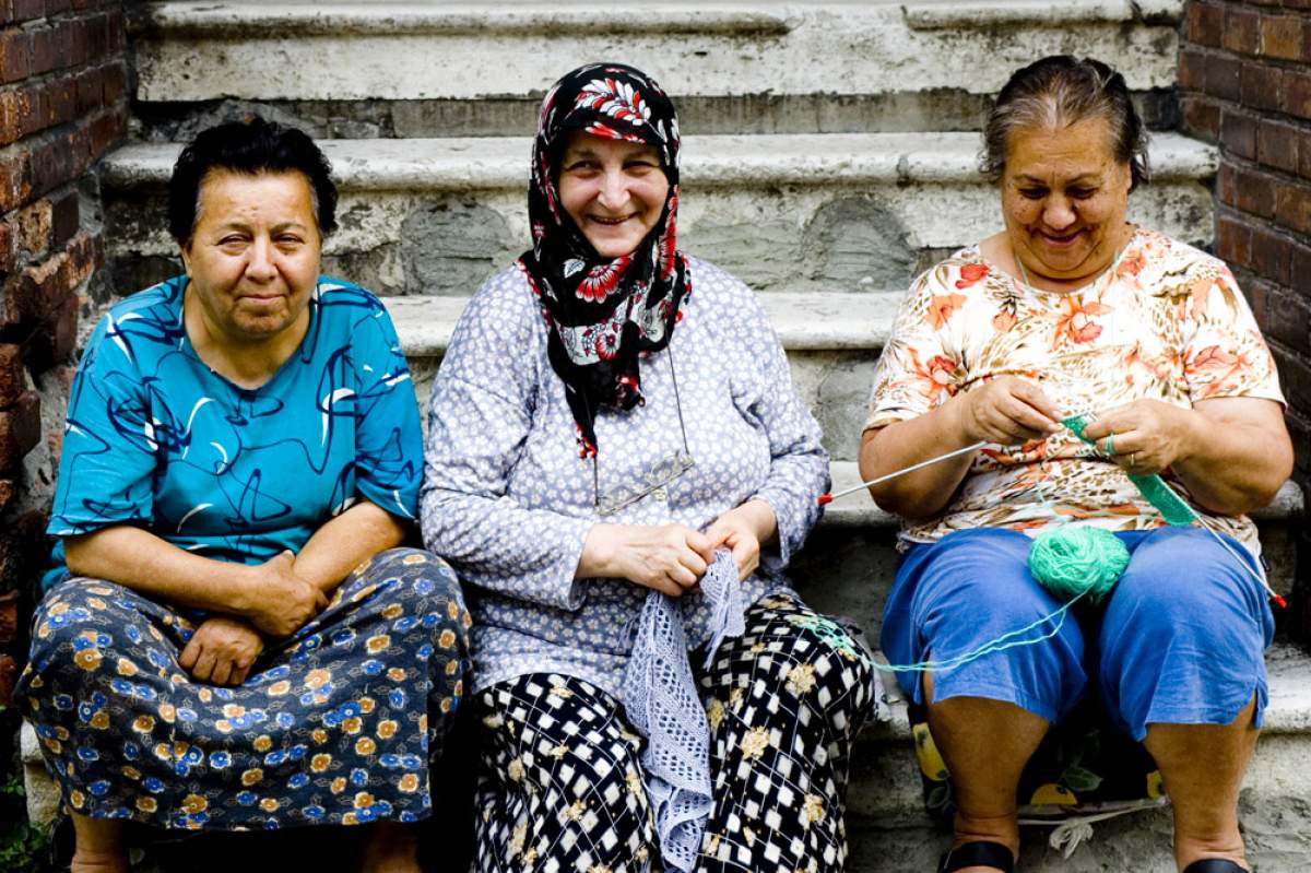Three old women are chilling on a  stoop. The woman on the left has very short curly hair and is dressed in blue. The middle old woman is wearing a headscarf and smiling into the camera. The one on the furthest right is wearing orange and crocheting a blanket.