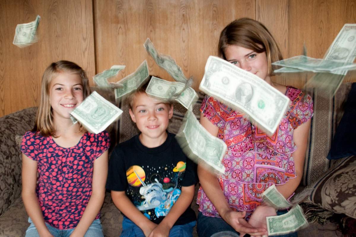Three Caucasian children are sitting on a couch. In the foreground are dollar bills that it appears they have thrown.