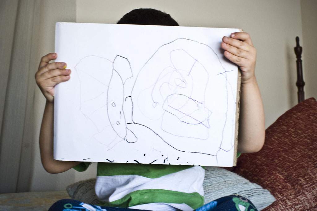 A child holding up a sideways image of a picture he has drawn. The child's face can't be seen. The drawing appears to be of a child or a blobby Prince Leia from Star Wars.