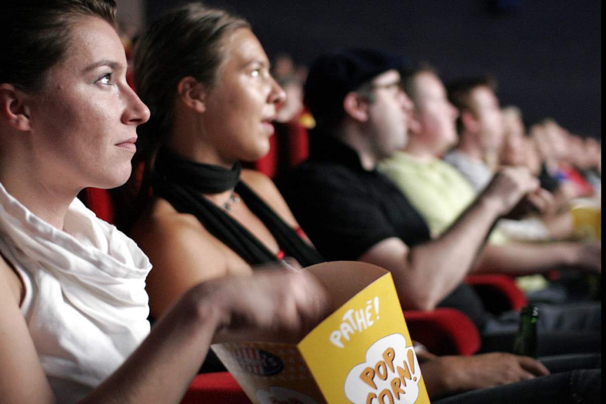 On the left side of the screen, two women are in focus watching a movie. The woman closest to the camera has her hand in a yellow popcorn tub.