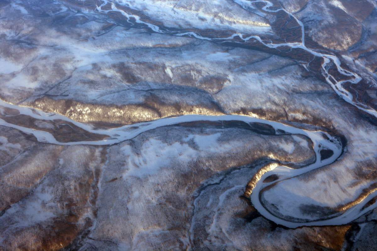 A photograph taken of Siberia from an airplane window. The ground appears to be mostly white ice and brown dirt. There is a large swirl in the center that the photographer identifies as an unknown river.