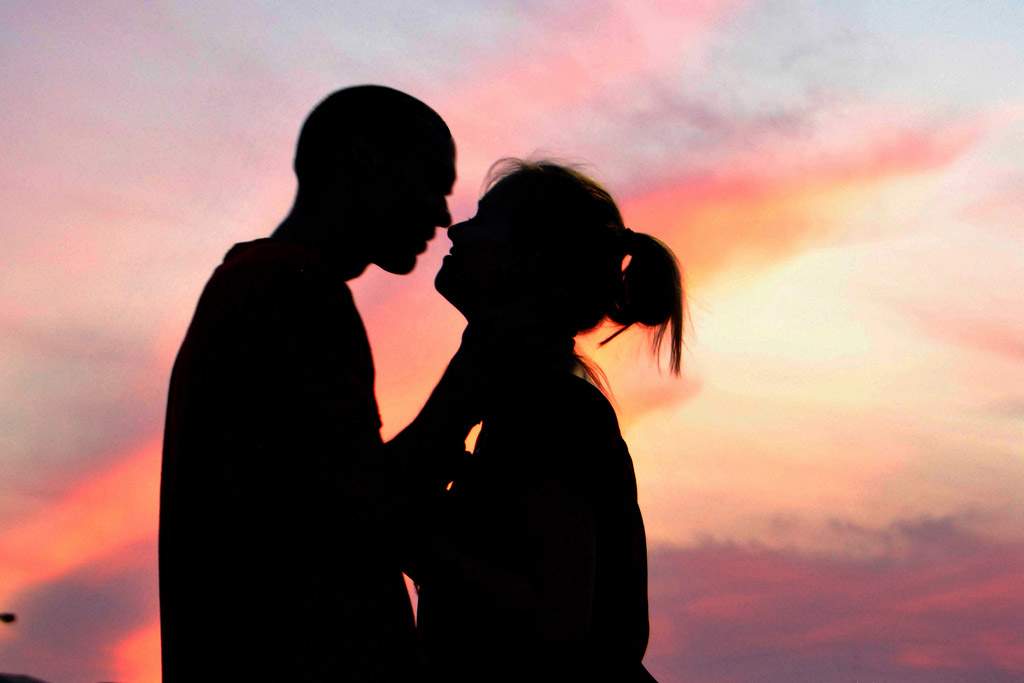A couple embraces in front of a sunset. Because of the lighting, they appear only as dark shadows against the blue, orange, pink, and purple of the sunset.