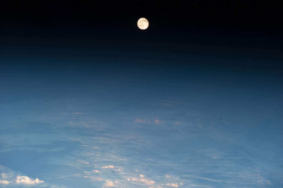 A waning gibbous moon over eastern Europe. The moon is a bright white circle in the very middle of the top of the image. Below the moon,  there are are light blue clouds and at the very bottom of the image, white clouds.