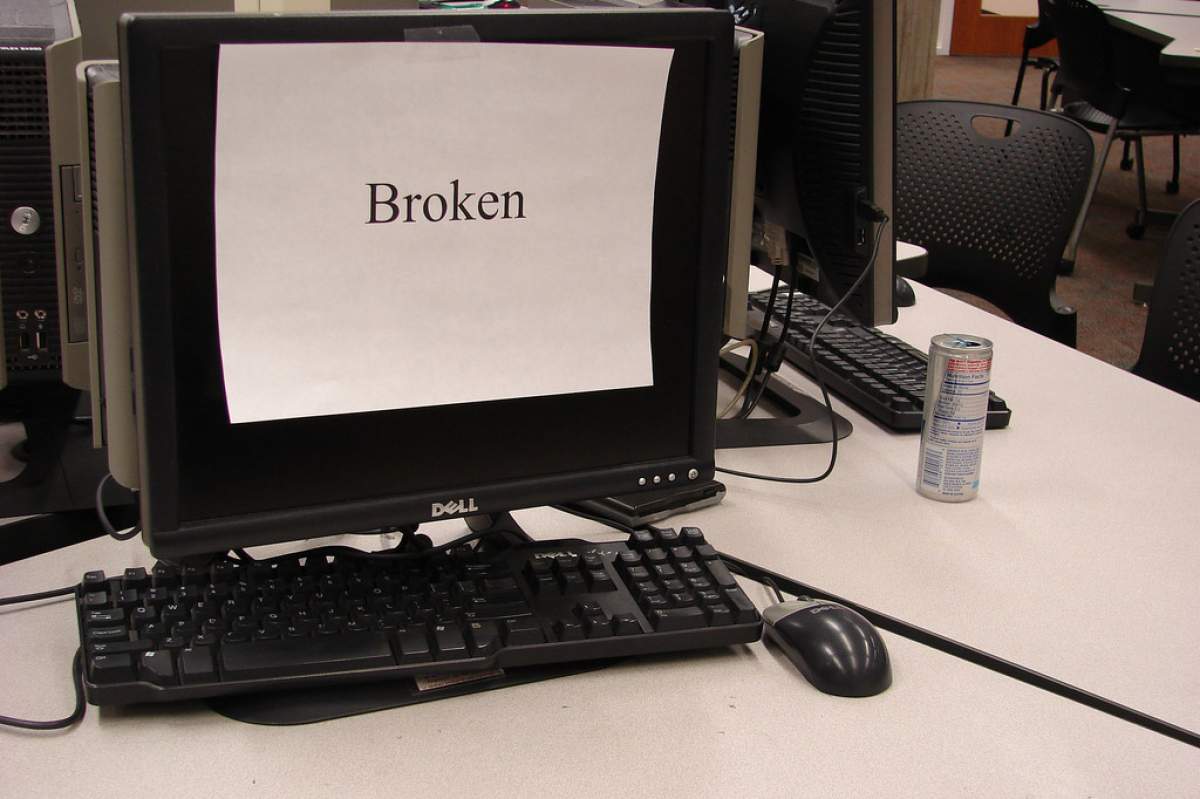 A PC computer (monitor, mouse, and keyboard all black). The screen is covered by a white sheet of paper with one word on it: Broken. All of this is on a white table. Next to it is an open can of what appears to be Red Bull.