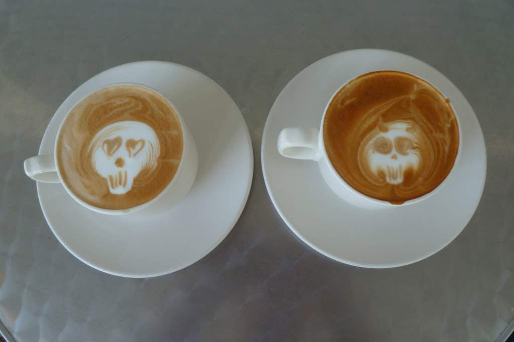 An overhead shot of two white coffee cups. The coffee cup on the left has white foam art in the shape of a skull. The skull's eyes are hearts. It's in the center of the cup with a light brown coffee background. The cup on the right also has  white skull as foam art. But its eyes are flat brown circles. The background is a darker brown.