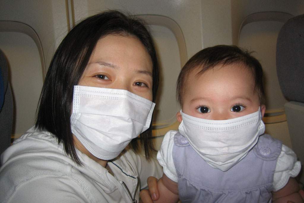 A woman and baby are facing the camera. They appear to be on an airplane. Both are wearing white surgical masks.