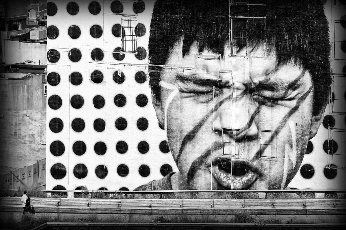A black and white image. The largest part of this image is a man's face making a pained expression with three marks running from his dark hair across his right eye, nose, and mouth. The background of this is black dots on a white background. This image is also a billboard. In the left bottom corner, small people are looking at the face.