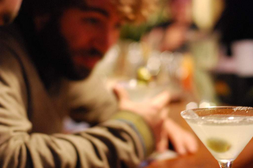 In the lower right corner of the image is an in focus martini with a single olive at the bottom of the glass. The rest of the image is blurry: a man with a beard wearing an olive green shirt is looking toward the martini. In the background are blurred shapes that look like they could be people standing up and a bar.
