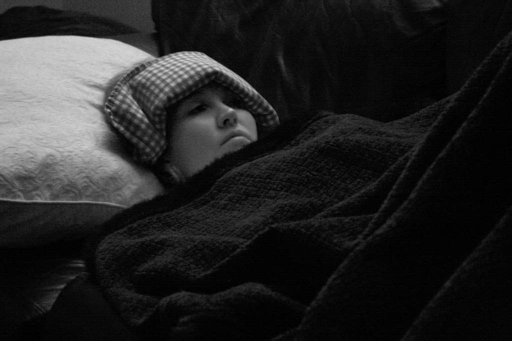 A black and white photo. A woman is in bed, her head on a white pillow, a heating pad with a gingham pattern is draped across her forehead. She has an unhappy look on her face. A black blanket that blends with the background is pulled up to her chin.