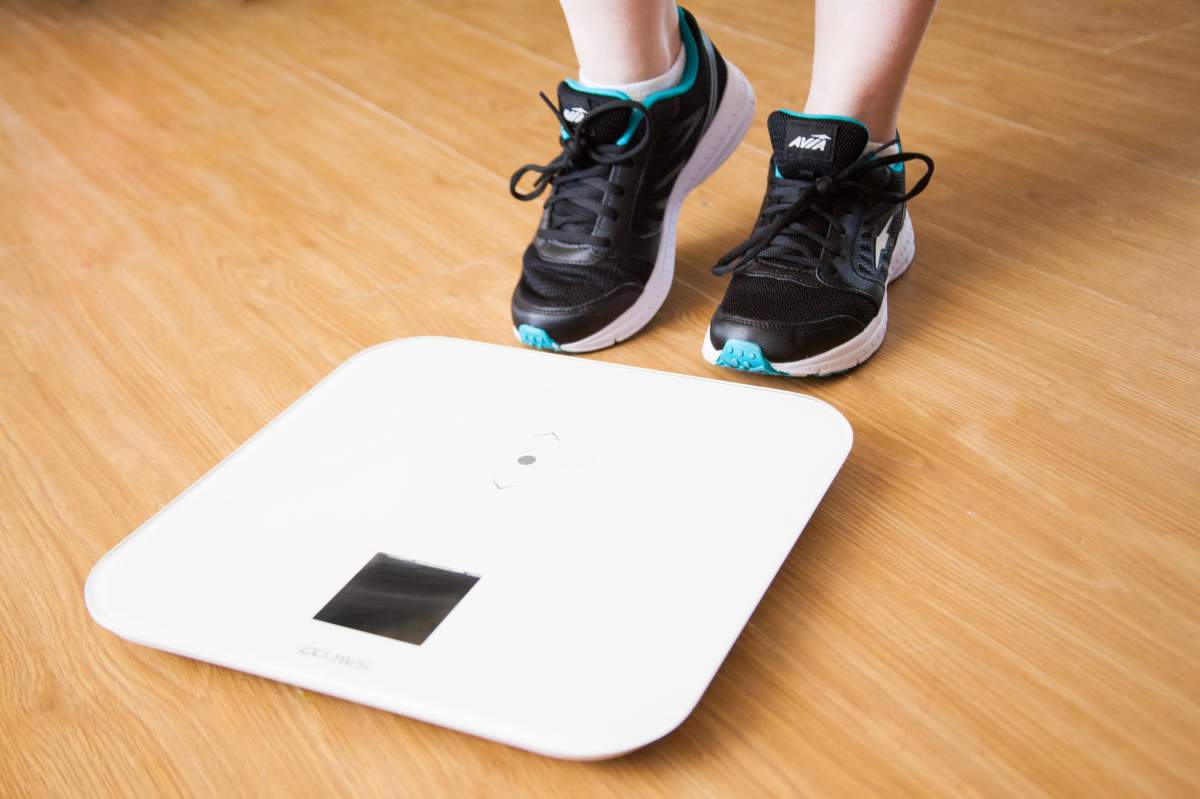A person wearing sneakers about to step on a white digital scale. The scale is on a hard wood floor.