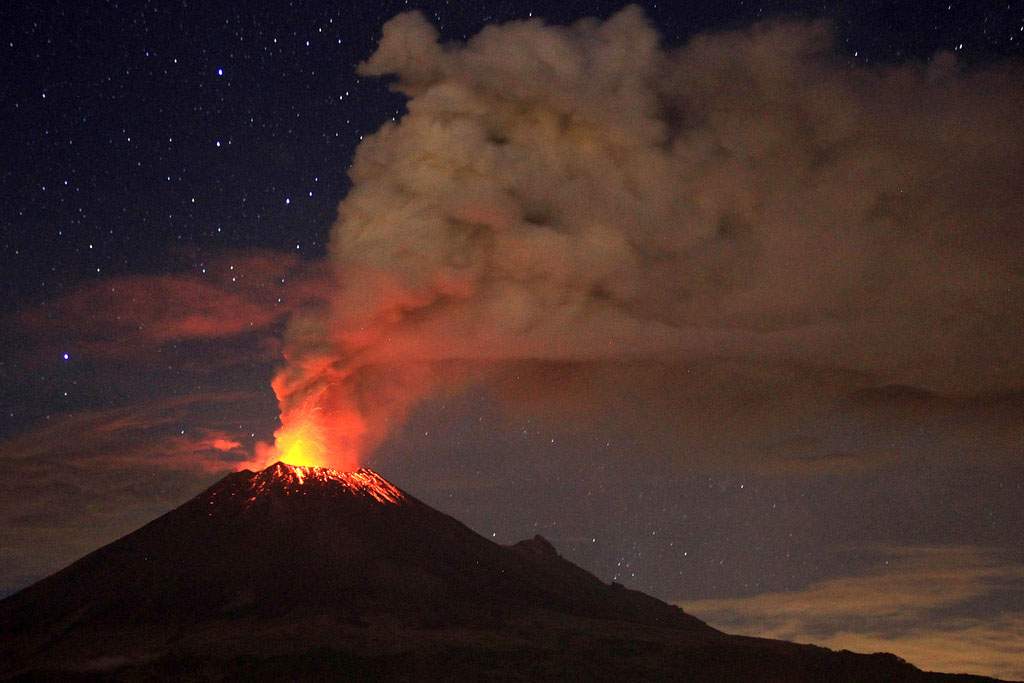 A volcano erupting in the night. The volcano is dark, but it's tip is bright red with lava. Gray smoke billows into the sky. In the background, night sky, stars, and clouds tinged pink from the lava's reflected light are visible.