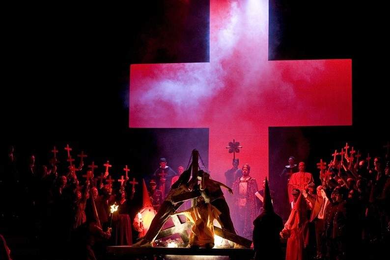 A crowd on stage appear to burn a heretic while a large red cross looms in the background