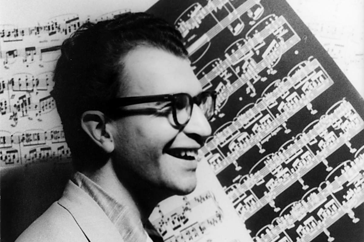 In black and white, a man in glasses smiles in front of sheet music