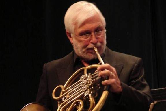 A bearded man in a black suit stands and plays the horn