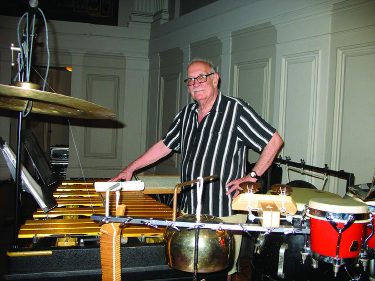 Man in striped shirt stands surrounded by percussion instruments.