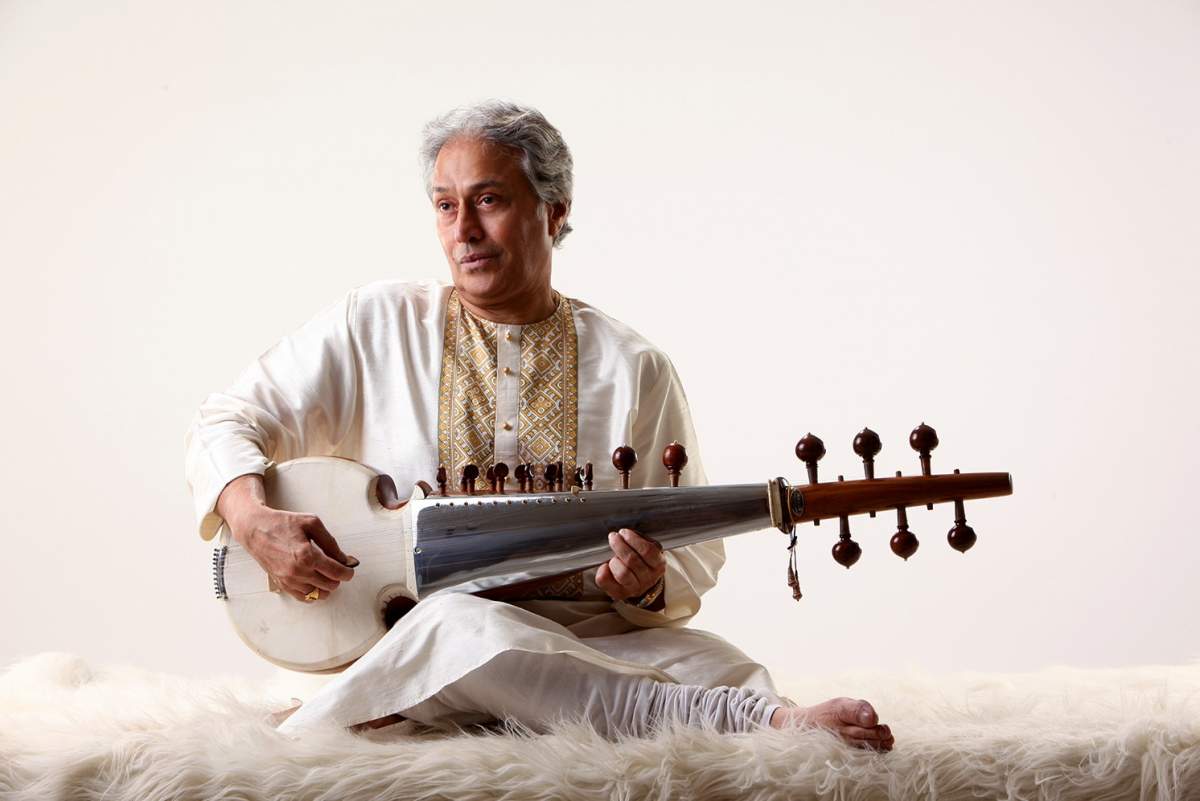 a man sits on a fur-lined surface playing a sarod