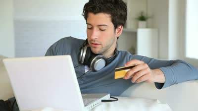 A young man wearing headphones holds a credit card an laptop - which indicates that he is buying music online.