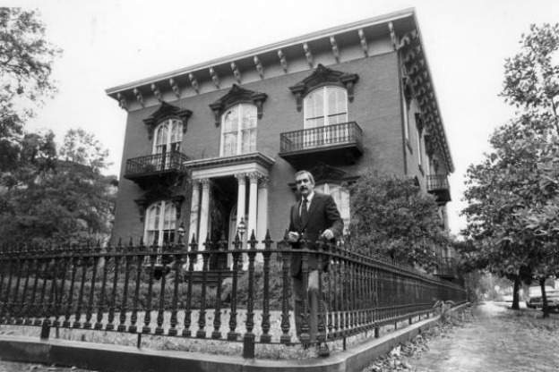 man in suit stands behind rod-iron fence in front of Italianate house in Savannah Georgia in this black-and-white photograph from 1980
