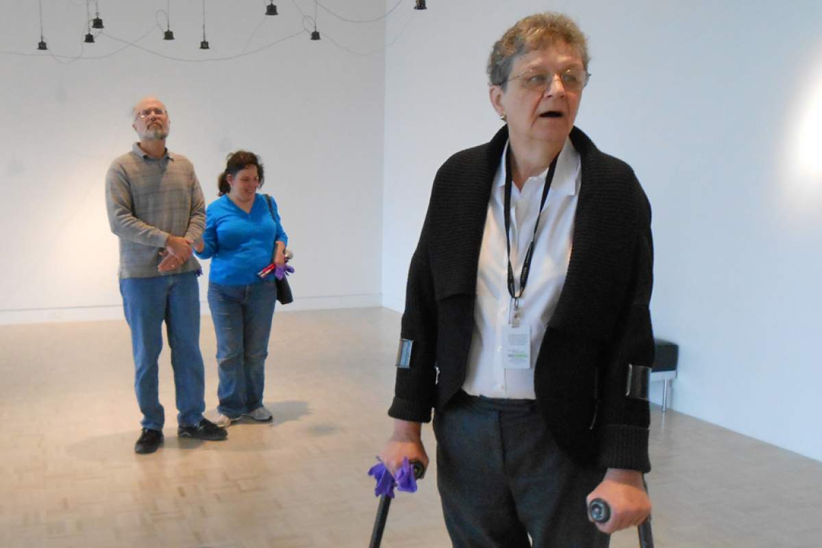 Marian Pettengill, using braces, leads a low-vision tour in gallery with white walls and ceiling at the Indianapolis Museum of Art. Art objects hang from the ceiling.  Tour participants Rhett Salisbury and Mary Stores stand in the background.
