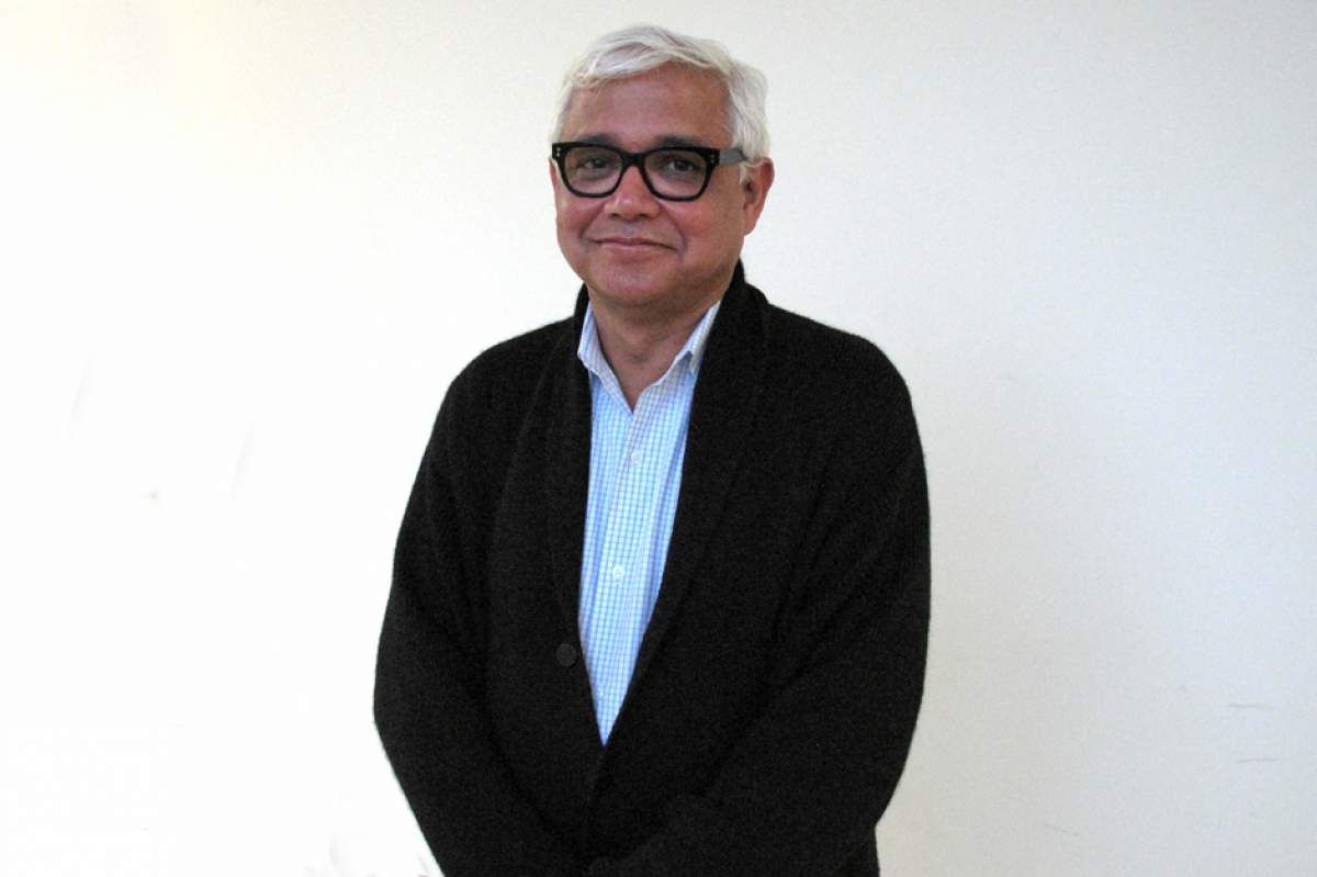 Amitav Ghosh with white hair, wearing black horn-rimmed glasses, open necked blue-and-white checked shirt, and dark gray cardigan sweater.