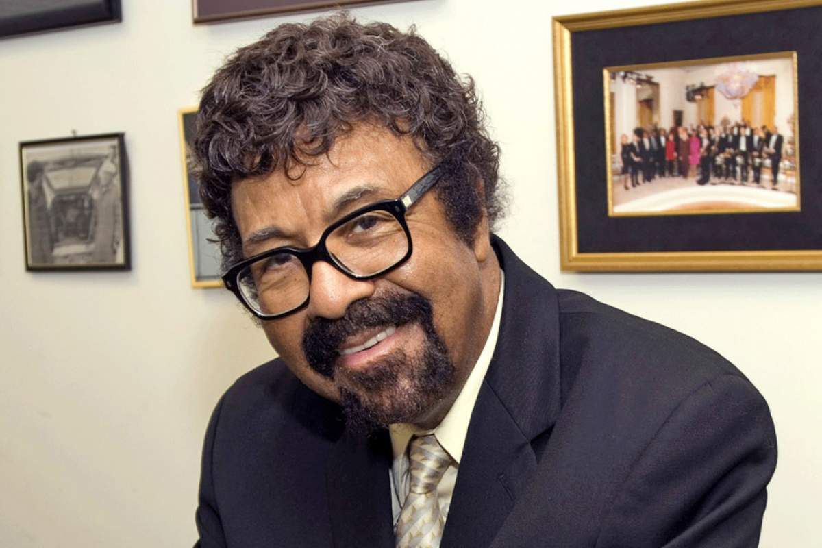 David Baker with framed pictures on wall behind him, wearing dark suit and tie, glasses, mustache and beard, smiling.