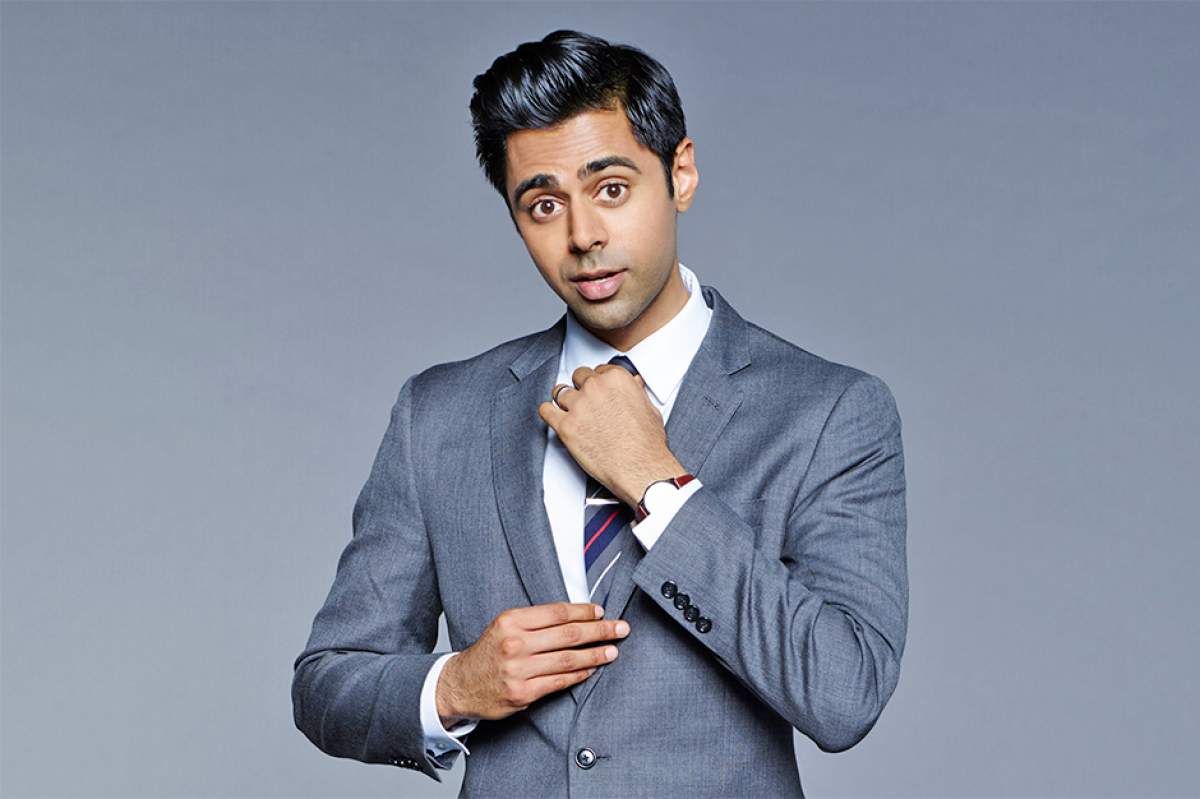Hasan Minhaj in gray suit, adjusting knot of his diagonally-striped necktie, looking at camera with quizzical-ironic expression.