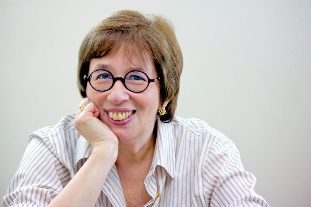 Linda Greenhouse, sitting with chin resting on right palm, wearing round-framed glasses, open-necked striped shirt, smiling.