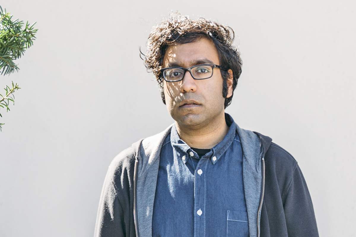 Hari Kondabolu outside in front of a white wall, wearing black horn-rimmed glasses, blue demin shirt open at neck, opened gray zippered sweatshirt, looking a bit confused.