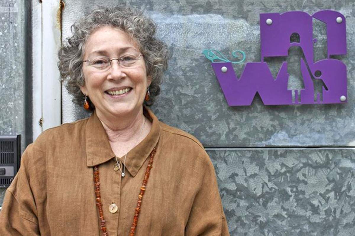 Toby Strout in wireframe glasses, salt-and-pepper hair, brown shirt and necklace, standing with purple stylized "Middle Way House" sign (stylized as MWH in purple letters) behind her.