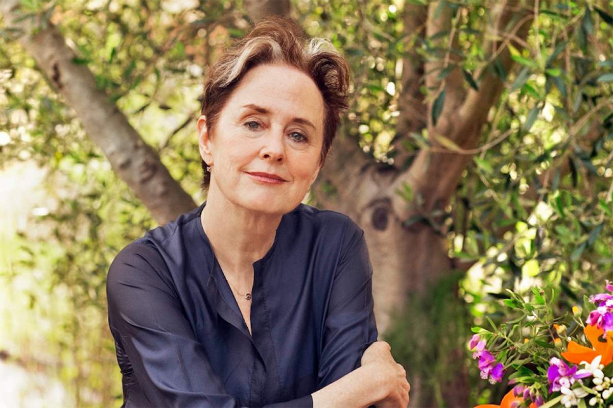 Alice Waters sitting outside wearing a silky purple top with trees in the background and flowers on the table.