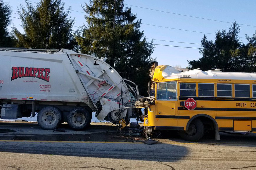 A yellow school bus is seen damaged right behind a rumpke garbage truck