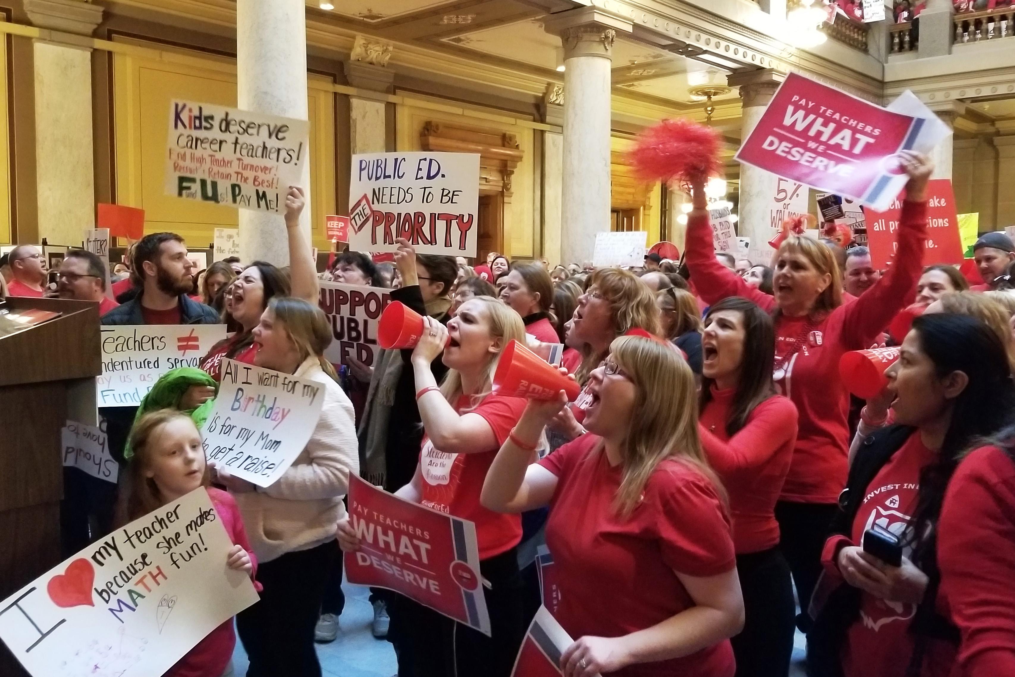 People in red shirts stand in the statehouse holding signs and chanting to support teacher pay